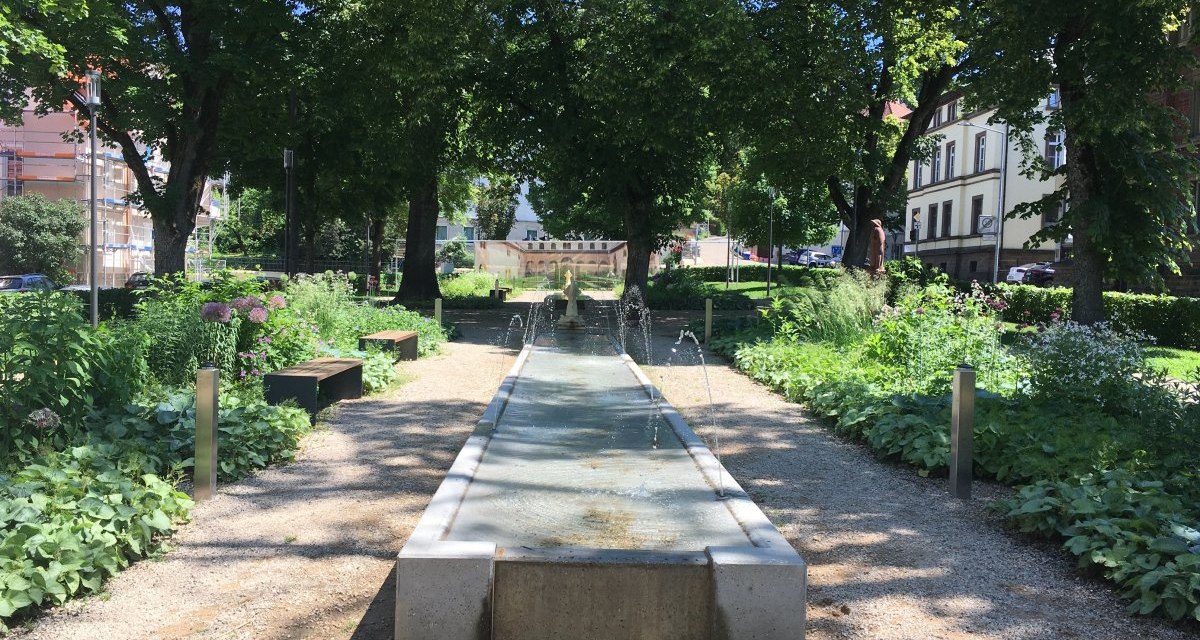 Ein schattiger Park mit einem langen Wasserbecken, umgeben von Bäumen, Bänken und Blumenbeeten., © Touristik und Marketing GmbH Ein schattiger Park mit einem langen Wasserbecken, umgeben von Bäumen, Bänken und Blumenbeeten., © Touristik und Marketing GmbH
