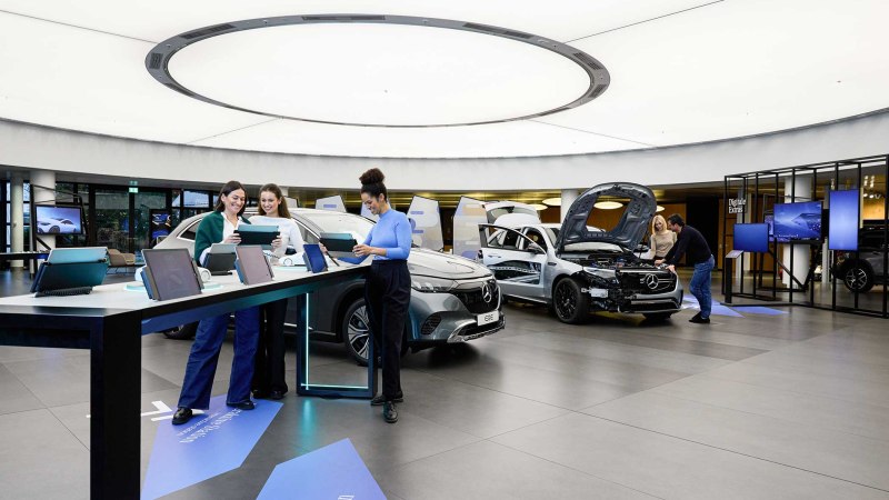 Moderner Ausstellungsraum mit Autos und interaktiven Stationen. Menschen nutzen Tablets, während andere ein Auto mit geöffneter Motorhaube betrachten., © Mercedes-Benz AG