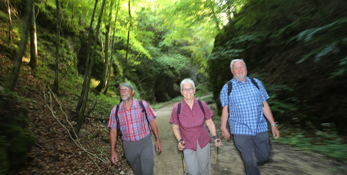 Drei ältere Personen wandern auf einem Waldweg in einer grünen Schlucht. Sie tragen karierte Hemden und Rucksäcke., © Bad Urach Tourismus