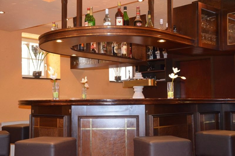 Elegante Hotelbar mit dunkler Holztheke, Flaschen im Regal und dekorativen Blumenvasen. Sitzhocker um die Theke verteilt., &copy; TOMAS