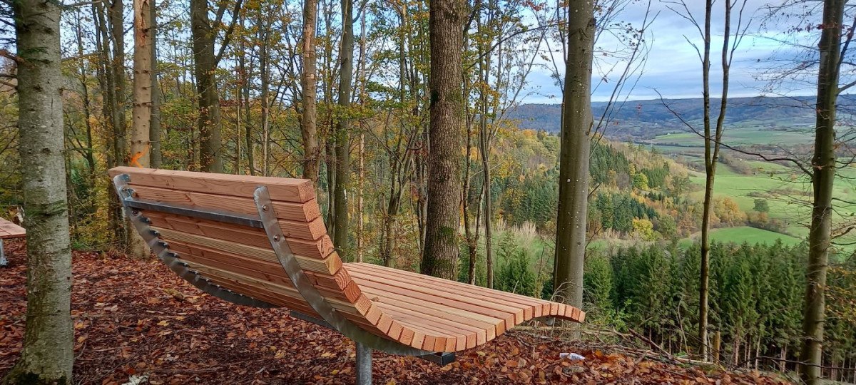 Holzbank am Aussichtspunkt im Wald, umgeben von Bäumen. Im Hintergrund eine weite, grüne Landschaft unter bewölktem Himmel., © Stadt Gaildorf