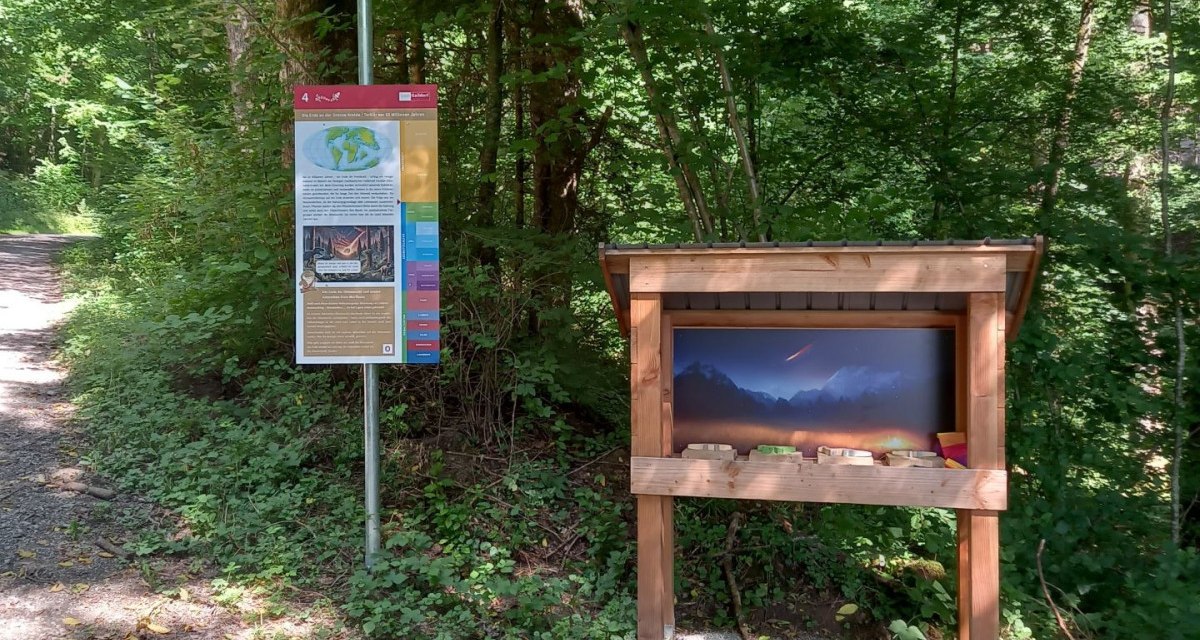 Eine Informationstafel und eine Wurfbude stehen im Wald auf dem Geopfad Gaildorf, Station 4. Die Tafel enthält geologische Informationen.