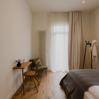 Minimalistisch eingerichtetes Schlafzimmer mit Holzboden, Bett, kleinem Schreibtisch und Stuhl. Vorhänge und ein Bademantel auf einem Ständer ergänzen das Bild., © Peter Oliver Wolff / DAS EDITH Minimalistisch eingerichtetes Schlafzimmer mit Holzboden, Bett, kleinem Schreibtisch und Stuhl. Vorhänge und ein Bademantel auf einem Ständer ergänzen das Bild., © Peter Oliver Wolff / DAS EDITH