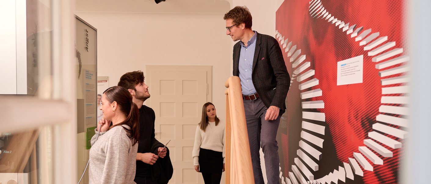 Menschen in einem Ausstellungsraum. Eine Person steht auf einer Treppe, andere betrachten eine Wand mit rotem Design. Thema: Angst., © Stiftung Psyche Menschen in einem Ausstellungsraum. Eine Person steht auf einer Treppe, andere betrachten eine Wand mit rotem Design. Thema: Angst., © Stiftung Psyche