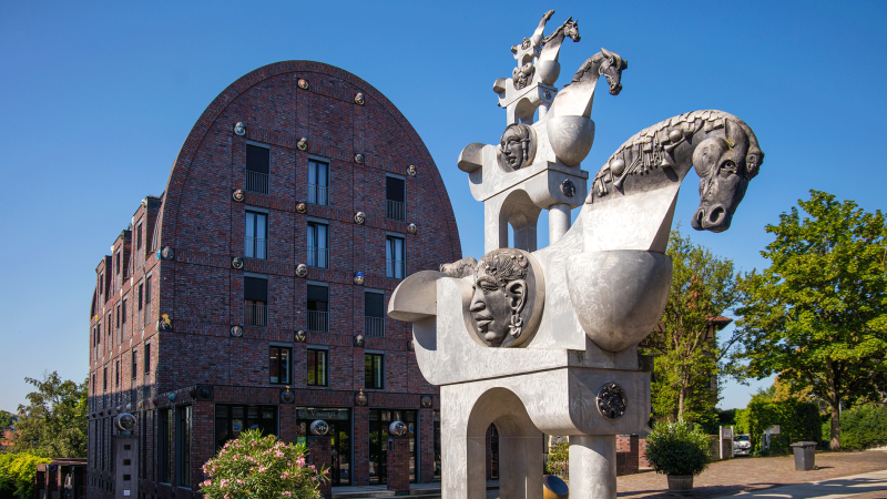 Moderne Skulptur mit Pferde- und Menschenköpfen vor einem Backsteingebäude mit rundem Dach, umgeben von Bäumen und Pflanzen., © Stuttgart-Marketing GmbH, Achim Mende Moderne Skulptur mit Pferde- und Menschenköpfen vor einem Backsteingebäude mit rundem Dach, umgeben von Bäumen und Pflanzen., © Stuttgart-Marketing GmbH, Achim Mende