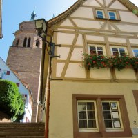 Fachwerkhaus mit blühendem Blumenkasten, daneben ein Kirchturm. Treppen führen zwischen den Gebäuden hinauf. Klarer blauer Himmel im Hintergrund., © Natur.Nah. Schönbuch & Heckengäu