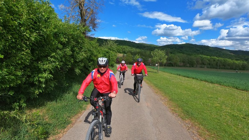 Drei Radfahrer in roten Jacken fahren auf einem l&auml;ndlichen Weg. Die Umgebung ist gr&uuml;n, mit B&auml;umen und Feldern, unter einem blauen Himmel mit Wolken., &copy; Land der 1000 H&uuml;gel - Kraichgau-Stromberg