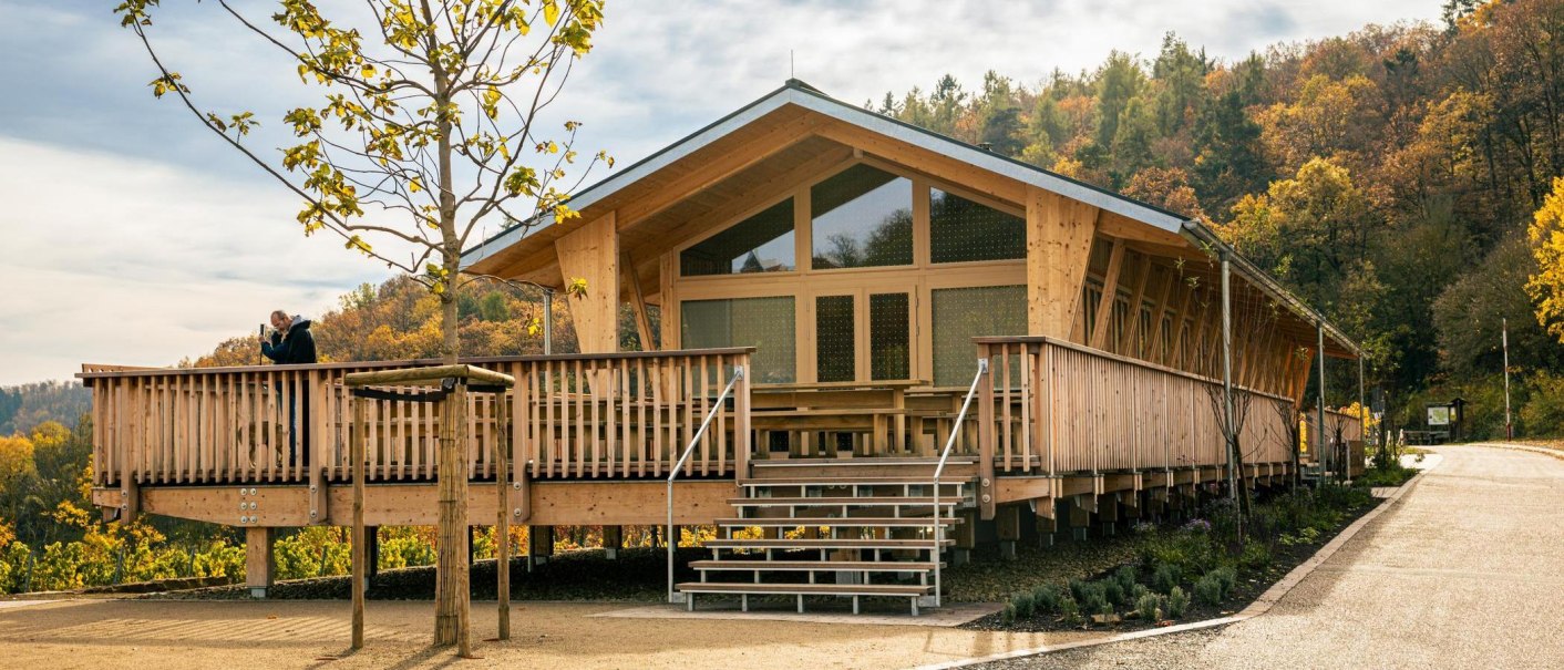 Ein modernes Holzgebäude mit großer Veranda in einer herbstlichen Landschaft. Ein Mann steht auf der Veranda und schaut in die Ferne., © Stuttgart-Marketing GmbH, Sarah Schmid