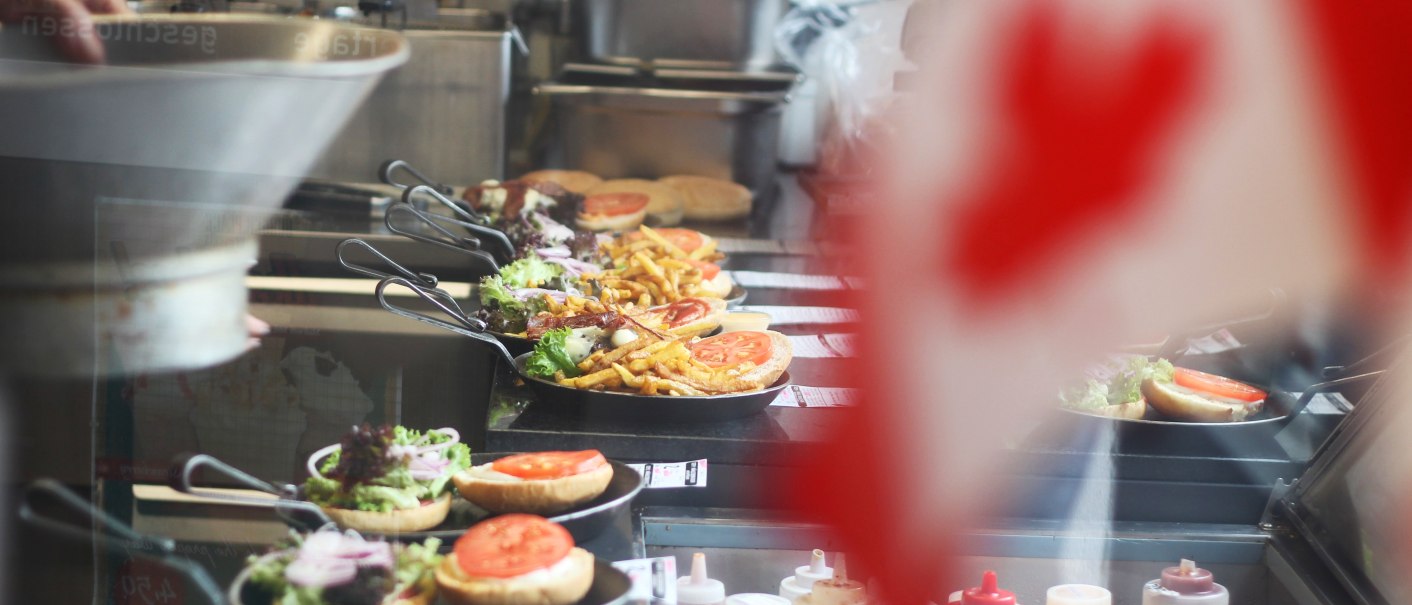 In einer Küche werden Burger und Pommes in Pfannen zubereitet. Im Vordergrund ist unscharf eine kanadische Flagge zu sehen., © SMG