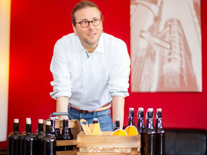 Ein Mann mit Brille präsentiert verschiedene Bierflaschen und eine Holzkiste mit Orangen vor einem roten Hintergrund., © PlochingenInfo Ein Mann mit Brille präsentiert verschiedene Bierflaschen und eine Holzkiste mit Orangen vor einem roten Hintergrund., © PlochingenInfo