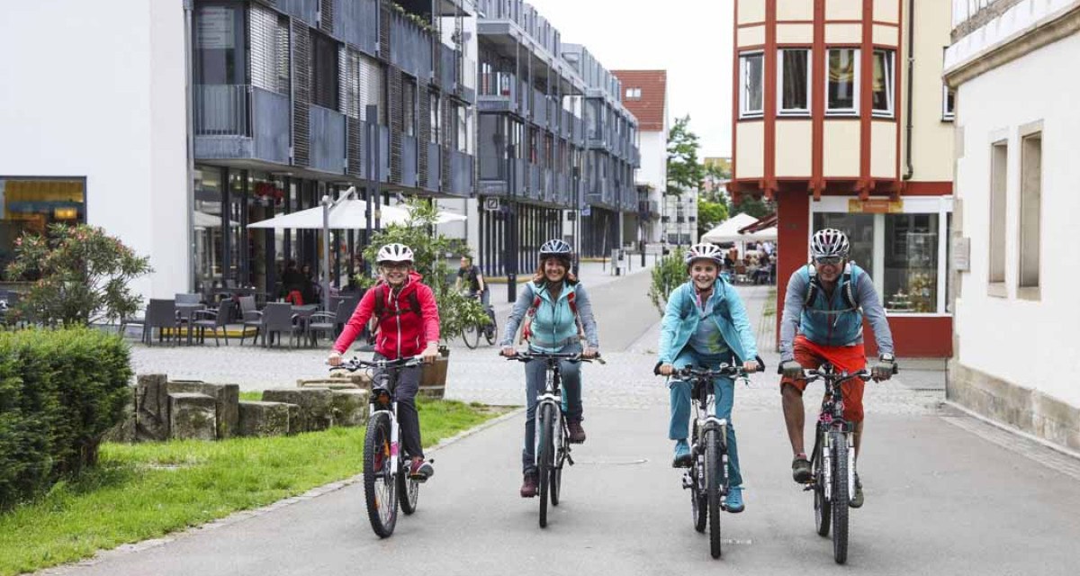 Vier Personen fahren mit Fahrrädern durch eine moderne Straße in Kirchheim unter Teck. Sie tragen Helme und Freizeitkleidung., © Schwäbische Alb Tourismusverband e.V. Vier Personen fahren mit Fahrrädern durch eine moderne Straße in Kirchheim unter Teck. Sie tragen Helme und Freizeitkleidung., © Schwäbische Alb Tourismusverband e.V.