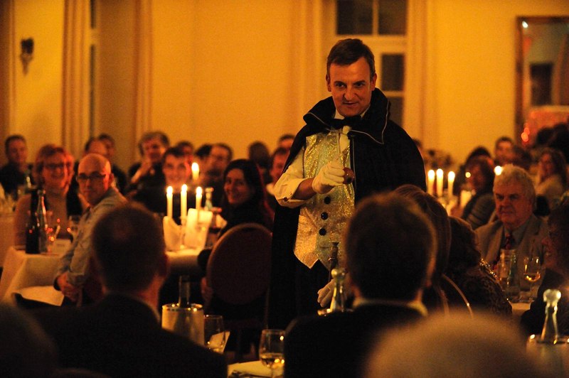 Ein Mann in einem viktorianischen Kostüm spricht zu einem Publikum bei einem Dinner-Event. Der Raum ist in warmes Kerzenlicht getaucht., © Gruseldinner GmbH & Co. KG Ein Mann in einem viktorianischen Kostüm spricht zu einem Publikum bei einem Dinner-Event. Der Raum ist in warmes Kerzenlicht getaucht., © Gruseldinner GmbH & Co. KG