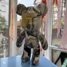 Bunte B&auml;renskulptur mit Pouringtechnik auf blauem Podest vor einem Fenster. Im Hintergrund sind bunte F&auml;den und ein Fachwerkhaus zu sehen., &copy; links im Bild