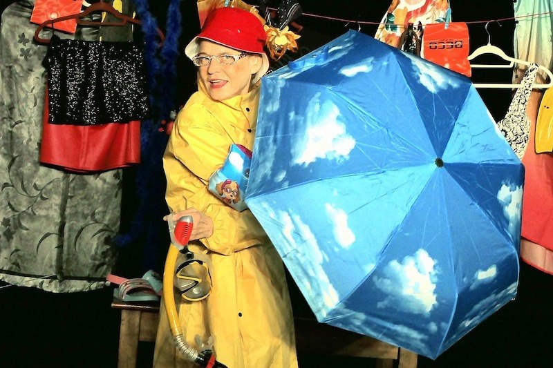 Eine Person in einem gelben Regenmantel und rotem Hut h&auml;lt einen blauen Regenschirm mit Wolkenmuster. Im Hintergrund h&auml;ngen bunte Kleidungsst&uuml;cke., &copy; Galli Theater Backnang