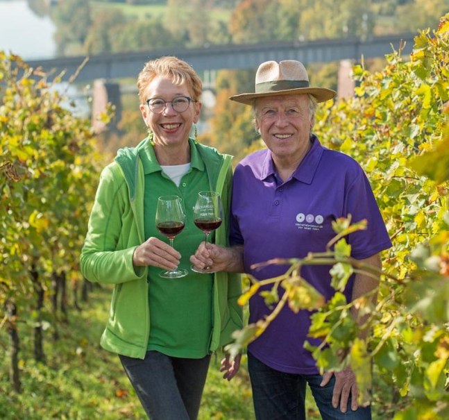 Zwei Personen genie&szlig;en Rotwein in einem sonnigen Weinberg. Sie l&auml;cheln und halten Weingl&auml;ser. Im Hintergrund ist eine Br&uuml;cke sichtbar., &copy; &copy; Hilde Hampp