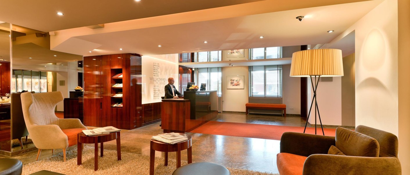 Elegante Hotel-Lobby mit Empfangstresen, gemütlichen Sesseln, Stehlampe und Teppich. Ein Mitarbeiter steht hinter dem Tresen., © Ringhotel Adler Asperg