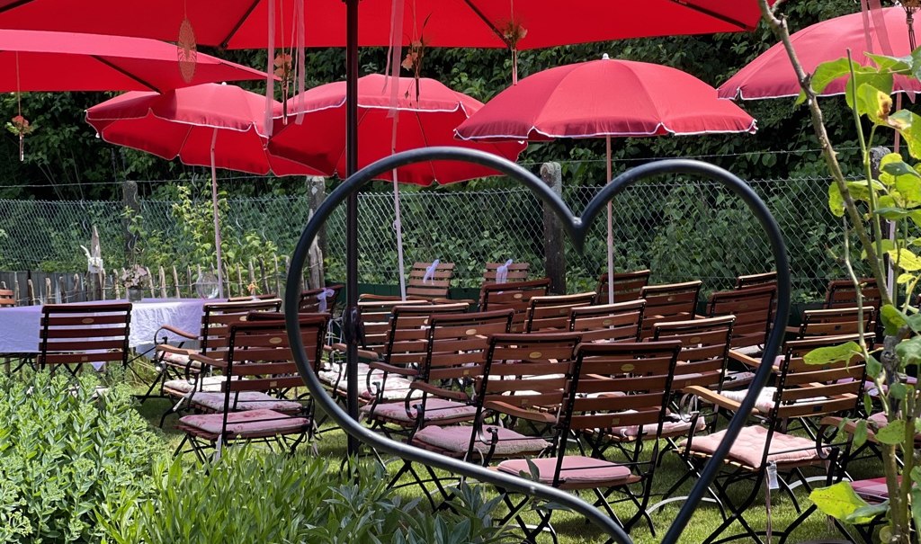 Ein Garten mit roten Sonnenschirmen und vielen Stühlen, eingerahmt von einem herzförmigen Metallrahmen. Ideal für Veranstaltungen im Freien., © Café Plätschwiesle