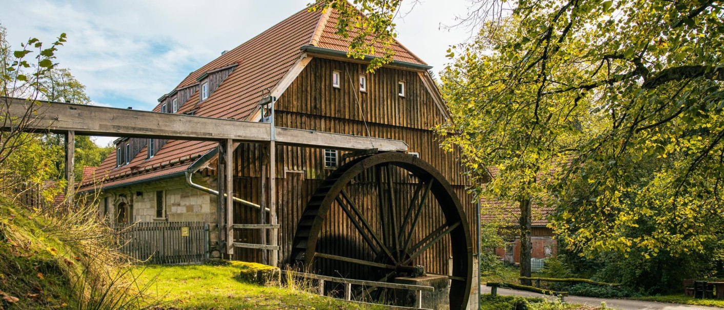 Eine historische Wassermühle mit großem Mühlrad, umgeben von Bäumen und Natur. Das Gebäude hat ein rotes Ziegeldach und Holzwände., © Stuttgart-Marketing GmbH, Sarah Schmid Eine historische Wassermühle mit großem Mühlrad, umgeben von Bäumen und Natur. Das Gebäude hat ein rotes Ziegeldach und Holzwände., © Stuttgart-Marketing GmbH, Sarah Schmid