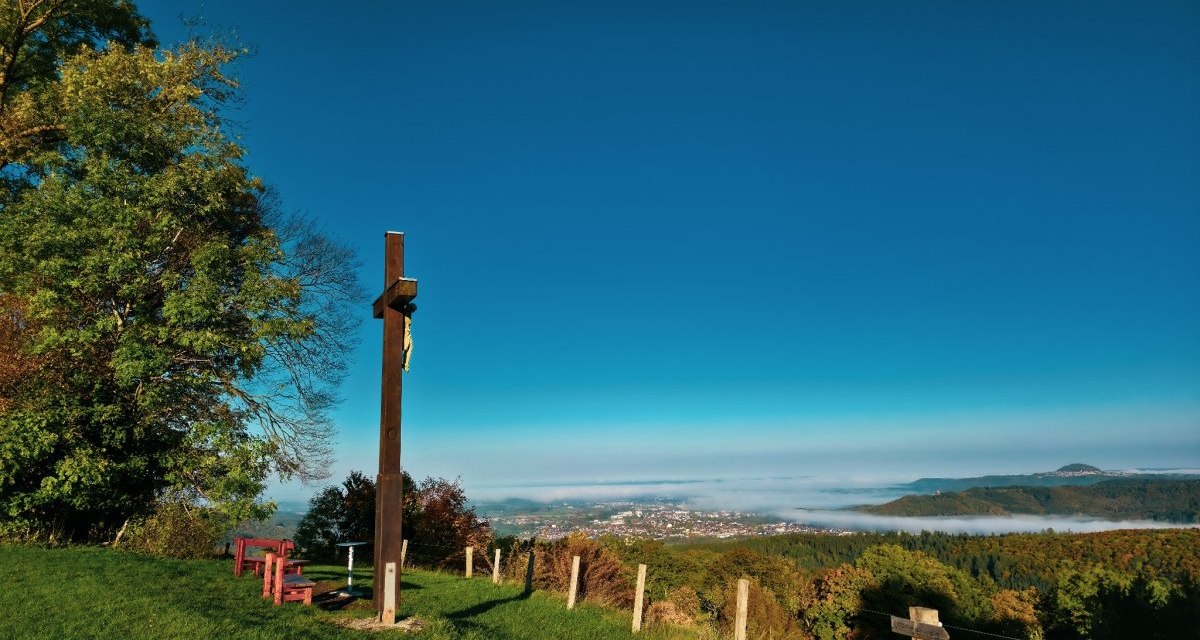Ein Holzkreuz steht auf einer grünen Wiese mit Blick auf ein nebelverhangenes Tal und eine Stadt in der Ferne. Der Himmel ist klar und blau., © Landkreis Göppingen Ein Holzkreuz steht auf einer grünen Wiese mit Blick auf ein nebelverhangenes Tal und eine Stadt in der Ferne. Der Himmel ist klar und blau., © Landkreis Göppingen