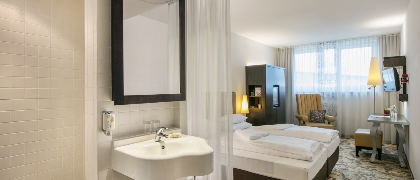 Ein modernes Hotelzimmer mit offenem Badezimmer, Doppelbett, Sessel, Schreibtisch und Fernseher. Helle Farben und stilvolle Einrichtung., &copy; ARCOTEL Hotels
