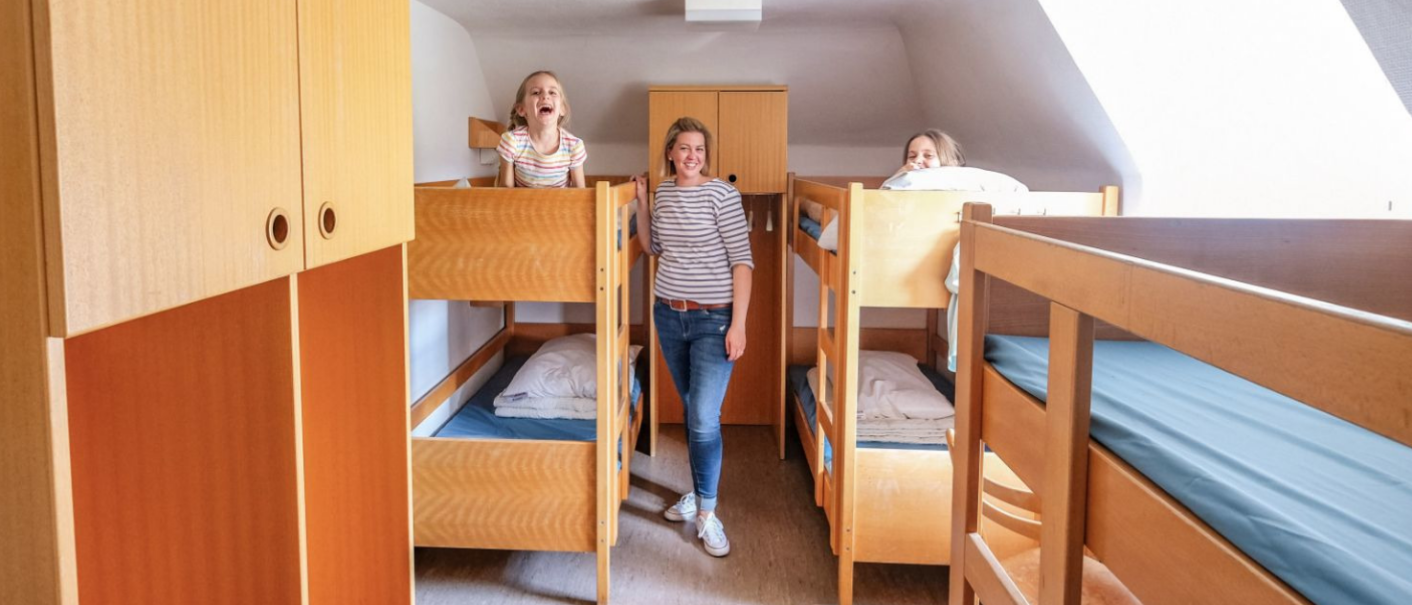 Ein Viererzimmer mit zwei Etagenbetten aus Holz. Drei Personen lächeln, zwei stehen auf den Betten, eine steht zwischen ihnen., © DJH Baden-Württemberg e.V.