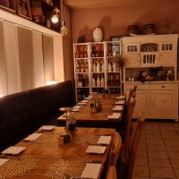 Ein gemütlicher Restaurantinnenraum mit gedeckten Holztischen, dekorativen Platzsets und warmem Licht. Regale mit Flaschen und ein rustikaler Schrank ergänzen das Ambiente., © RistoRocco Ein gemütlicher Restaurantinnenraum mit gedeckten Holztischen, dekorativen Platzsets und warmem Licht. Regale mit Flaschen und ein rustikaler Schrank ergänzen das Ambiente., © RistoRocco