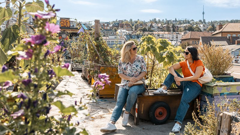 Zwei Frauen sitzen in einem urbanen Dachgarten mit Pflanzen und genießen den Ausblick auf die Stadt im Hintergrund., © © Stuttgart-Marketing GmbH, Sarah Schmid