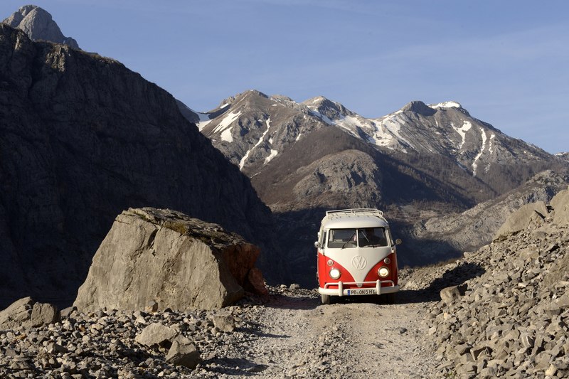 Ein roter VW Bulli auf einem steinigen Bergweg, umgeben von schneebedeckten Bergen unter klarem Himmel., © BLICKFANG Ein roter VW Bulli auf einem steinigen Bergweg, umgeben von schneebedeckten Bergen unter klarem Himmel., © BLICKFANG