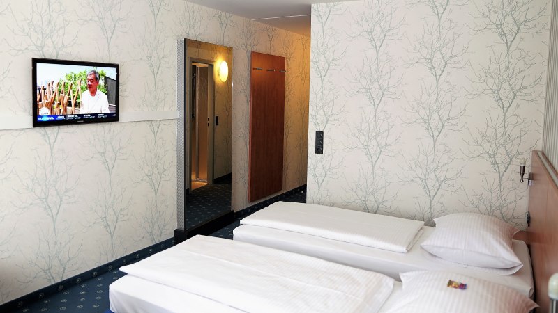 Hotelzimmer mit zwei Einzelbetten, einem Fernseher an der Wand und einem großen Spiegel. Die Wände sind mit einem baumartigen Muster tapeziert., © TOMAS Hotelzimmer mit zwei Einzelbetten, einem Fernseher an der Wand und einem großen Spiegel. Die Wände sind mit einem baumartigen Muster tapeziert., © TOMAS