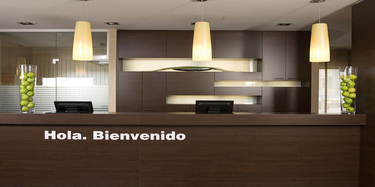 Moderne Rezeption mit dunklen Holzmöbeln, zwei Computern und Schriftzug 'Hola. Bienvenido'. Zwei Vasen mit grünen Äpfeln als Dekoration., © NH Ludwigsburg
