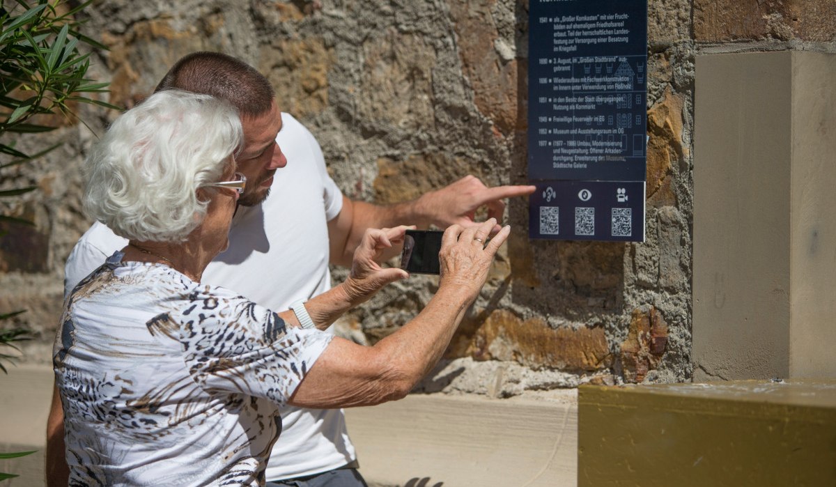 Zwei Personen betrachten eine Infotafel am Kornhaus. Eine ältere Frau scannt mit ihrem Smartphone einen QR-Code, während ein Mann auf die Tafel zeigt., © Stadt Kirchheim unter Teck