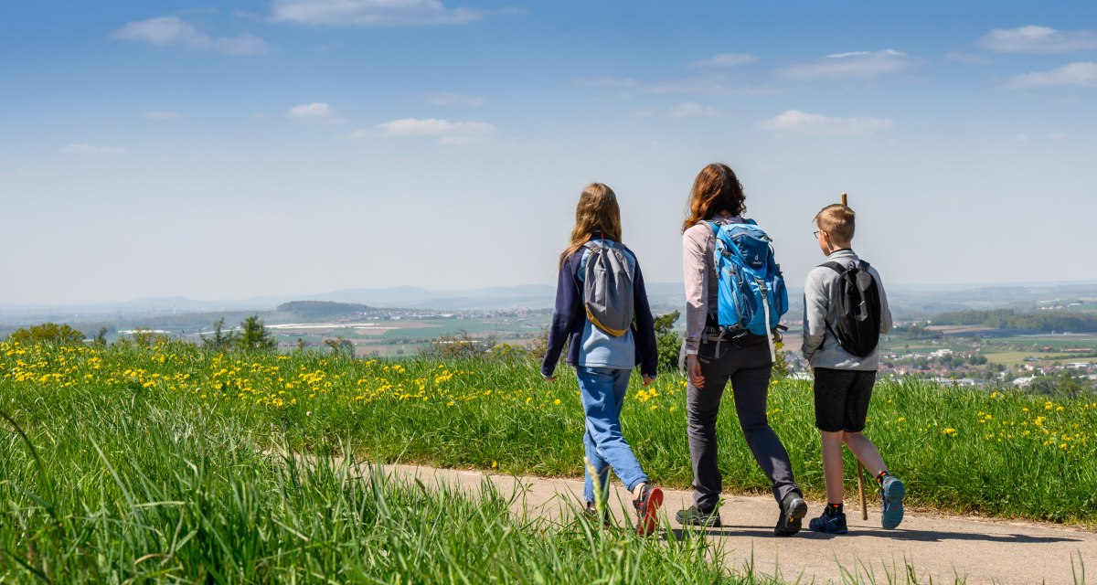 Drei Personen wandern auf einem Weg durch eine grüne Wiese mit gelben Blumen, im Hintergrund eine weite Landschaft unter blauem Himmel., © Stadtverwaltung Winnenden Drei Personen wandern auf einem Weg durch eine grüne Wiese mit gelben Blumen, im Hintergrund eine weite Landschaft unter blauem Himmel., © Stadtverwaltung Winnenden