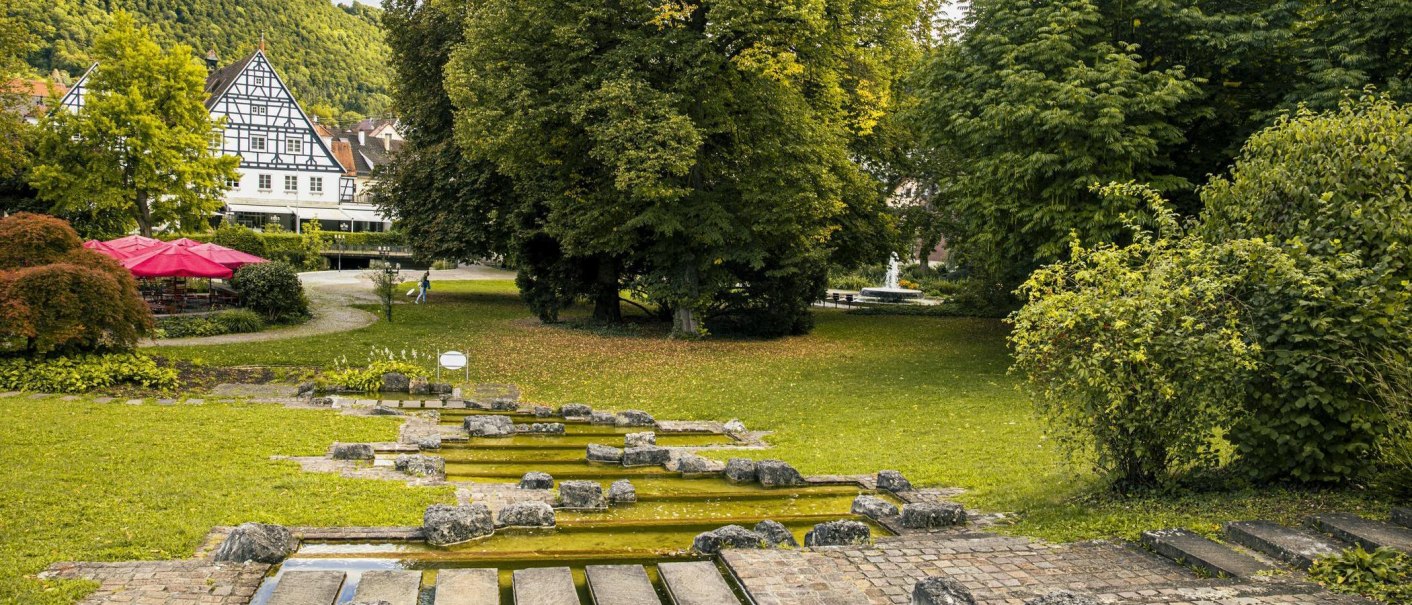 Grüner Kurpark in Bad Überkingen mit Fachwerkhaus, Bäumen, Brunnen und roten Sonnenschirmen., © Stuttgart-Marketing GmbH, Sarah Schmid Grüner Kurpark in Bad Überkingen mit Fachwerkhaus, Bäumen, Brunnen und roten Sonnenschirmen., © Stuttgart-Marketing GmbH, Sarah Schmid