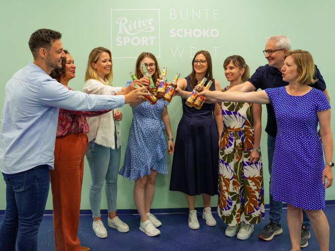 Eine Gruppe von Menschen st&ouml;&szlig;t mit bunten Schokoladenflaschen vor einer Wand mit Ritter Sport Logo an., &copy; Alfred Ritter GmbH &amp; Co. KG | Waldenbuch