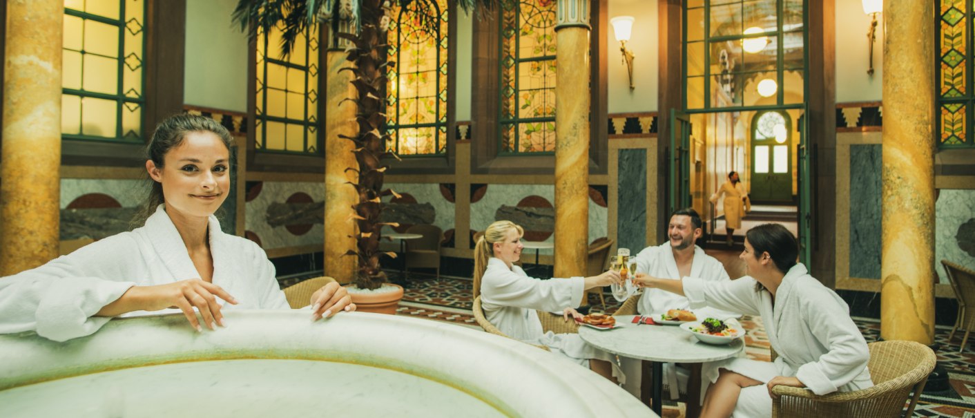 Personen in Badem&auml;nteln entspannen in einer eleganten Halle mit Palmen und Mosaikfenstern. Sie sto&szlig;en mit Getr&auml;nken an und genie&szlig;en Snacks., &copy; &copy;LOCHER Fotodesign & Manufaktur &copy;Staatsbad Wildbad GmbH