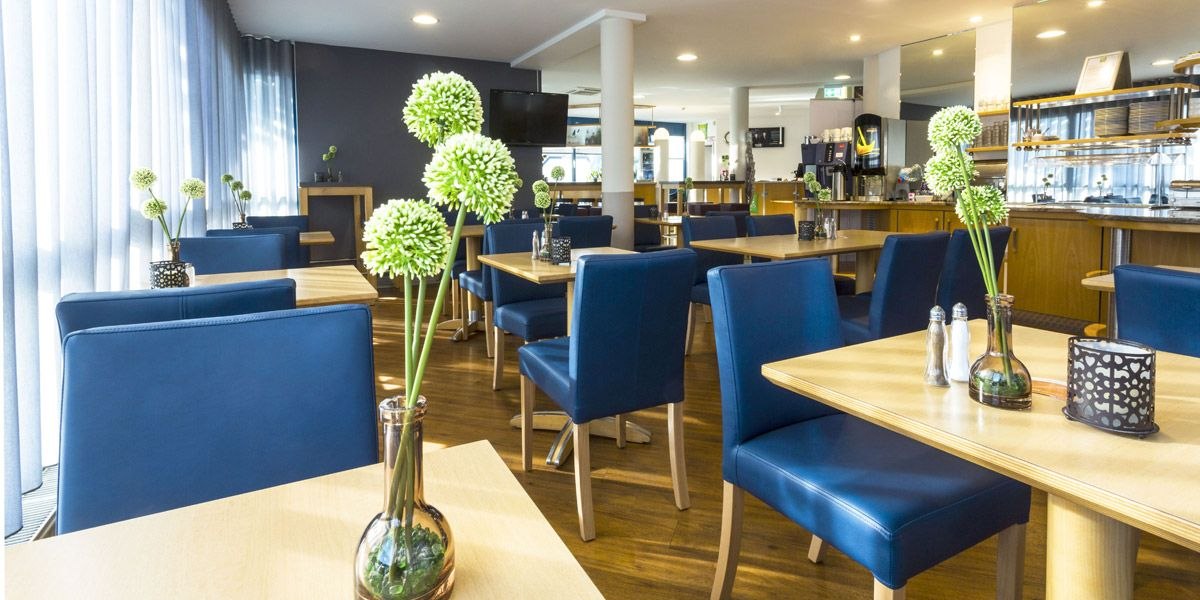 Ein modernes Restaurant mit blauen Stühlen, Holztischen und grünen Blumenvasen. Helle Beleuchtung und ein Buffetbereich im Hintergrund., © ibis Styles Filderstadt Messe Stuttgart Ein modernes Restaurant mit blauen Stühlen, Holztischen und grünen Blumenvasen. Helle Beleuchtung und ein Buffetbereich im Hintergrund., © ibis Styles Filderstadt Messe Stuttgart