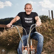 Mann auf Fahrrad mit "Dregg-Sagg" T-Shirt, lächelnd vor ländlicher Kulisse., © links im Bild