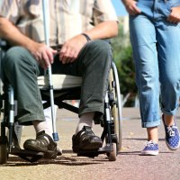 Ein Mann sitzt in einem Rollstuhl auf einem Gehweg, während eine Person in Jeans und Turnschuhen daneben geht., © pixabay