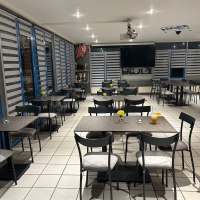 Ein modernes Bistro mit grauen Tischen und Stühlen, dekoriert mit gelben Blumen. Im Hintergrund sind Trophäen und ein großer Bildschirm zu sehen., © cBistro09 Ein modernes Bistro mit grauen Tischen und Stühlen, dekoriert mit gelben Blumen. Im Hintergrund sind Trophäen und ein großer Bildschirm zu sehen., © cBistro09