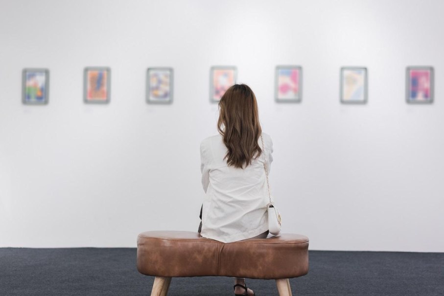 Eine Frau sitzt auf einer Bank in einer Galerie und betrachtet verschwommene Kunstwerke an der weißen Wand., © AdobeStock582052129 Kiattisak Eine Frau sitzt auf einer Bank in einer Galerie und betrachtet verschwommene Kunstwerke an der weißen Wand., © AdobeStock582052129 Kiattisak