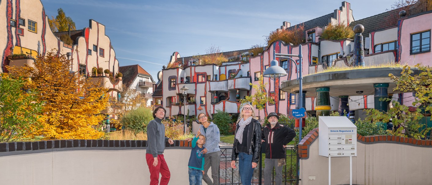 Menschen stehen vor dem farbenfrohen Hundertwasser-Wohnhaus in Plochingen. Die Fassade ist wellenförmig und bunt gestaltet, umgeben von Herbstbäumen., © Bildergalerie Attilla