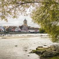 Weitwinkelaufnahme der Fischtreppe in Nürtingen. Im Vordergrund der Fluss mit Steinen, im Hintergrund historische Gebäude und ein Kirchturm, umrahmt von Bäumen., © SMG, Sarah Schmid