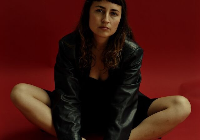 Eine Person in schwarzer Lederjacke sitzt mit verschränkten Beinen auf einem roten Hintergrund und schaut in die Kamera., © Stadt Böblingen - Amt für Kultur Eine Person in schwarzer Lederjacke sitzt mit verschränkten Beinen auf einem roten Hintergrund und schaut in die Kamera., © Stadt Böblingen - Amt für Kultur