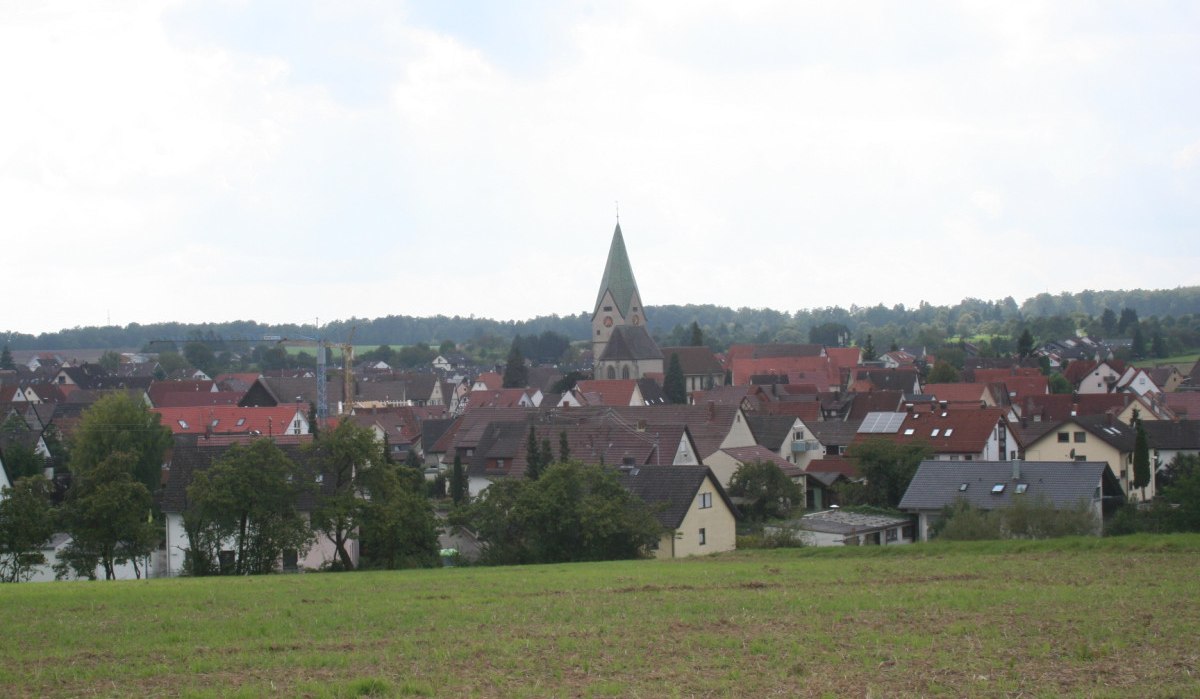 Dorfansicht mit Kirche und roten Dächern, umgeben von Bäumen und Feldern unter bewölktem Himmel., © Natur.Nah. Schönbuch & Heckengäu Dorfansicht mit Kirche und roten Dächern, umgeben von Bäumen und Feldern unter bewölktem Himmel., © Natur.Nah. Schönbuch & Heckengäu