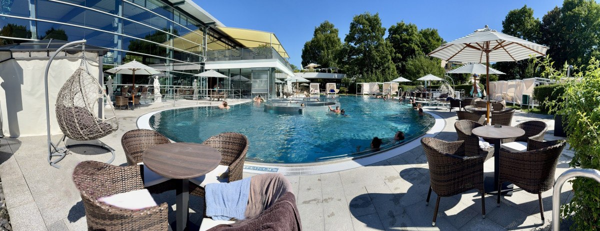 Ein modernes Au&szlig;enbecken mit Liegest&uuml;hlen und Sonnenschirmen. Menschen entspannen im Wasser und auf der Terrasse. Umgeben von B&auml;umen und einem modernen Geb&auml;ude., &copy; AlbThermen Bad Urach