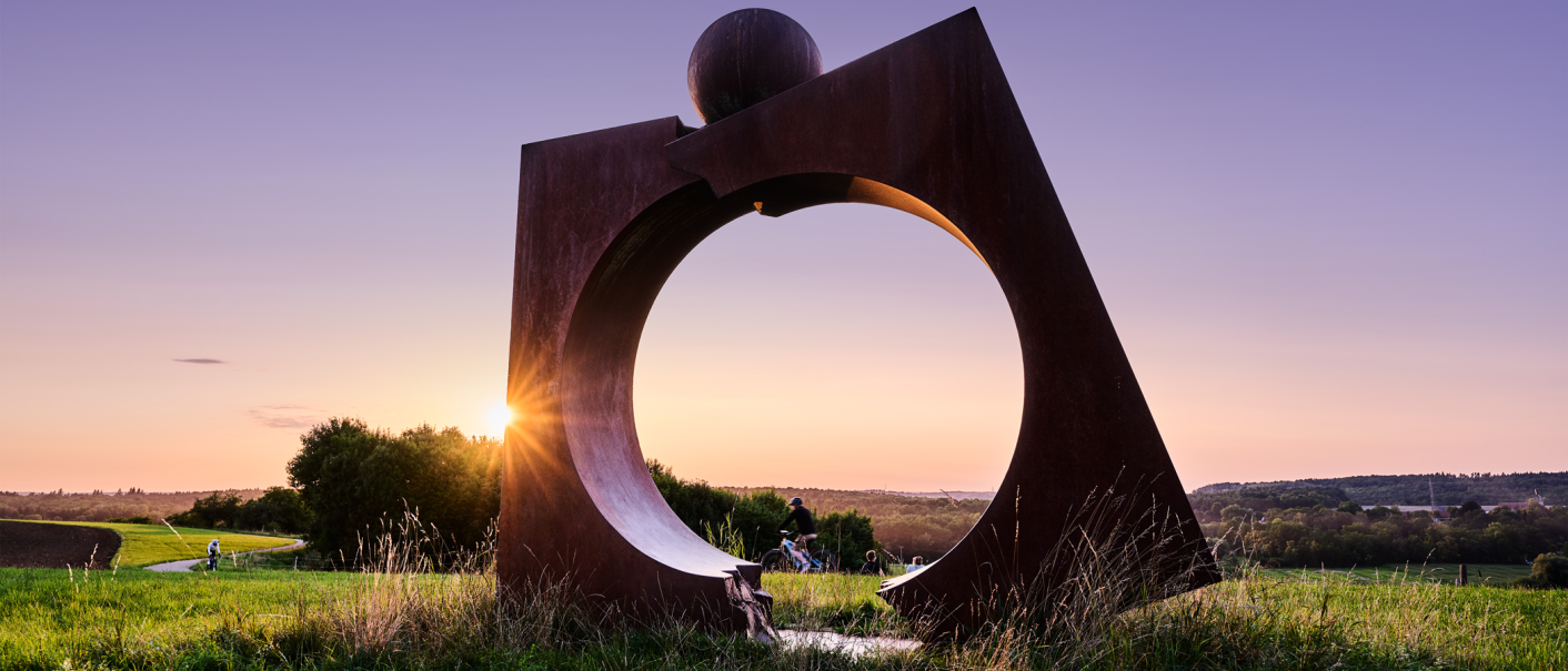 Eine große Metallskulptur steht auf einem Feld, während die Sonne am Horizont untergeht. Menschen spazieren und radeln im Hintergrund., © Andreas Sporn Eine große Metallskulptur steht auf einem Feld, während die Sonne am Horizont untergeht. Menschen spazieren und radeln im Hintergrund., © Andreas Sporn