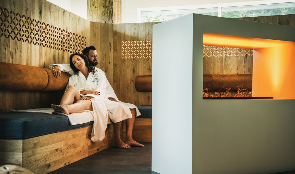 Ein Paar in Bademänteln entspannt in einer modernen Sauna mit Holzwänden und einem Kamin. Die Atmosphäre wirkt warm und gemütlich., © LOCHER Fotodesign&Manufaktur Ein Paar in Bademänteln entspannt in einer modernen Sauna mit Holzwänden und einem Kamin. Die Atmosphäre wirkt warm und gemütlich., © LOCHER Fotodesign&Manufaktur