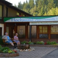 Geb&auml;ude mit Schild 'Hochwald Appartements' und 'Praxis f&uuml;r Physiotherapie EPPEL'. Zwei Personen sitzen auf einer Bank, umgeben von Pflanzen und B&auml;umen., &copy; Hotel Hochwald