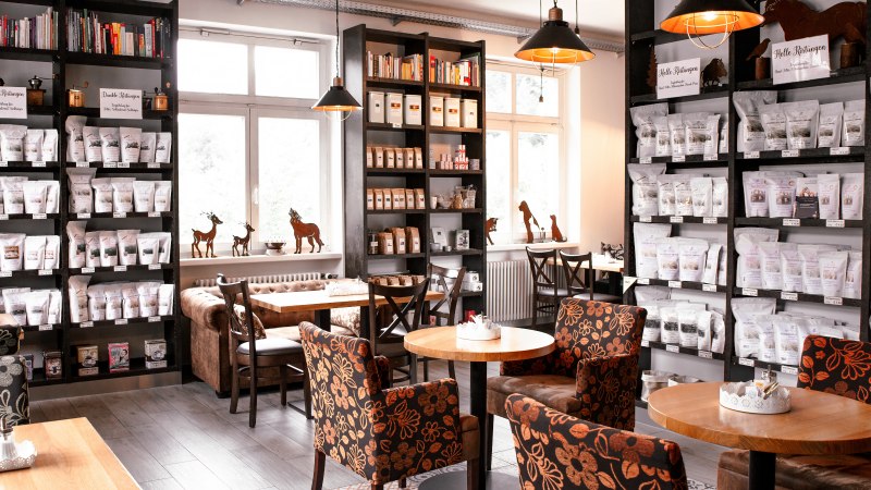 Gemütliches Café mit Regalen voller Kaffeepackungen, Tischen und floral gemusterten Sesseln., © Kaffee- Manufaktur Bad Wildbad, Jens Schröck Visionärsfabrik Keltern Gemütliches Café mit Regalen voller Kaffeepackungen, Tischen und floral gemusterten Sesseln., © Kaffee- Manufaktur Bad Wildbad, Jens Schröck Visionärsfabrik Keltern