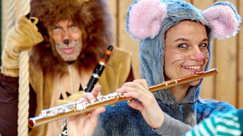 Zwei Personen in Tierkostümen, eine als Maus mit Flöte, die andere als Löwe mit Klarinette, vor einer Holzwand. Sie wirken fröhlich und kreativ., © Backnang Zwei Personen in Tierkostümen, eine als Maus mit Flöte, die andere als Löwe mit Klarinette, vor einer Holzwand. Sie wirken fröhlich und kreativ., © Backnang